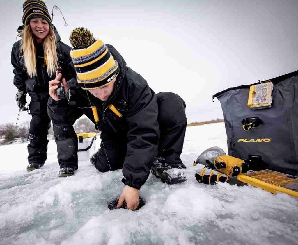 Man and woman ice fishing in black gear with ice fishing poles, lines, and tackle box.