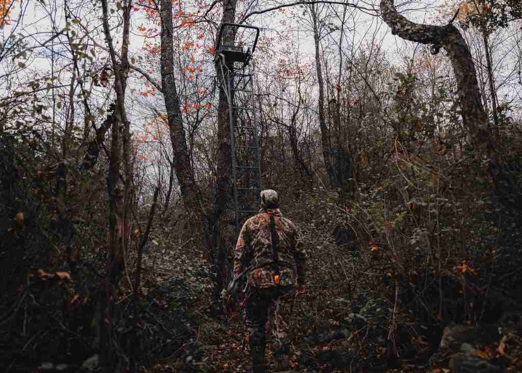 A hunter walks towards a tall ladder stand in the forest while wearing camouflage hunting garb