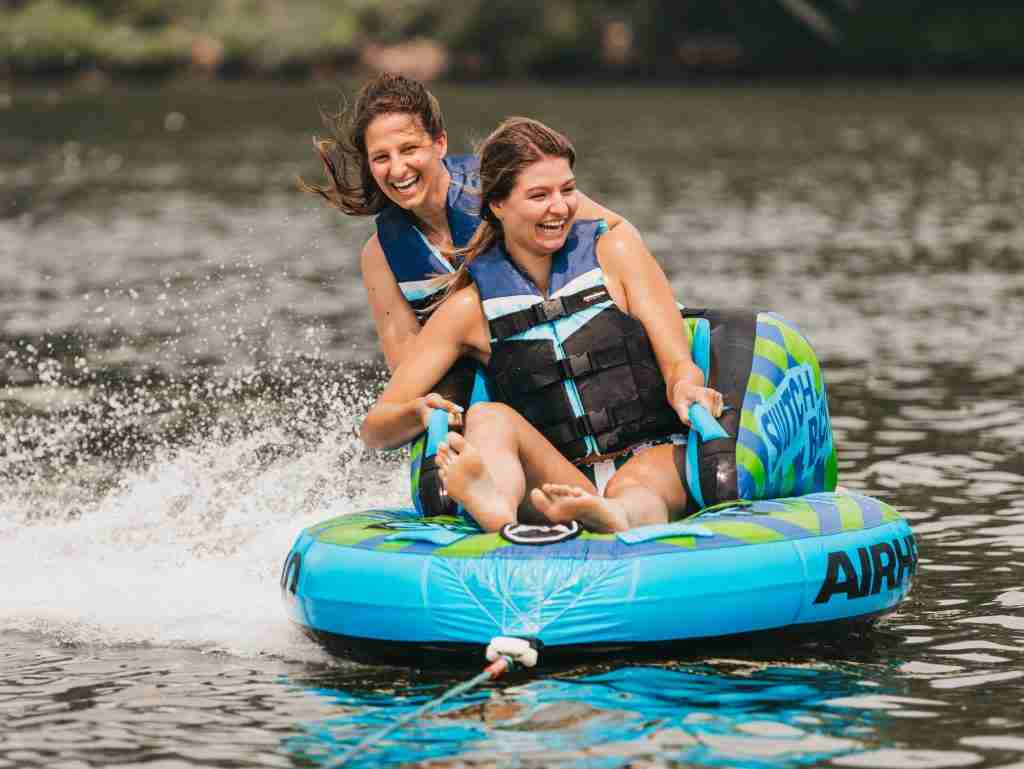 Two girls tubing on an inflatable tube pulled by a boat while wearing traditional life jackets
