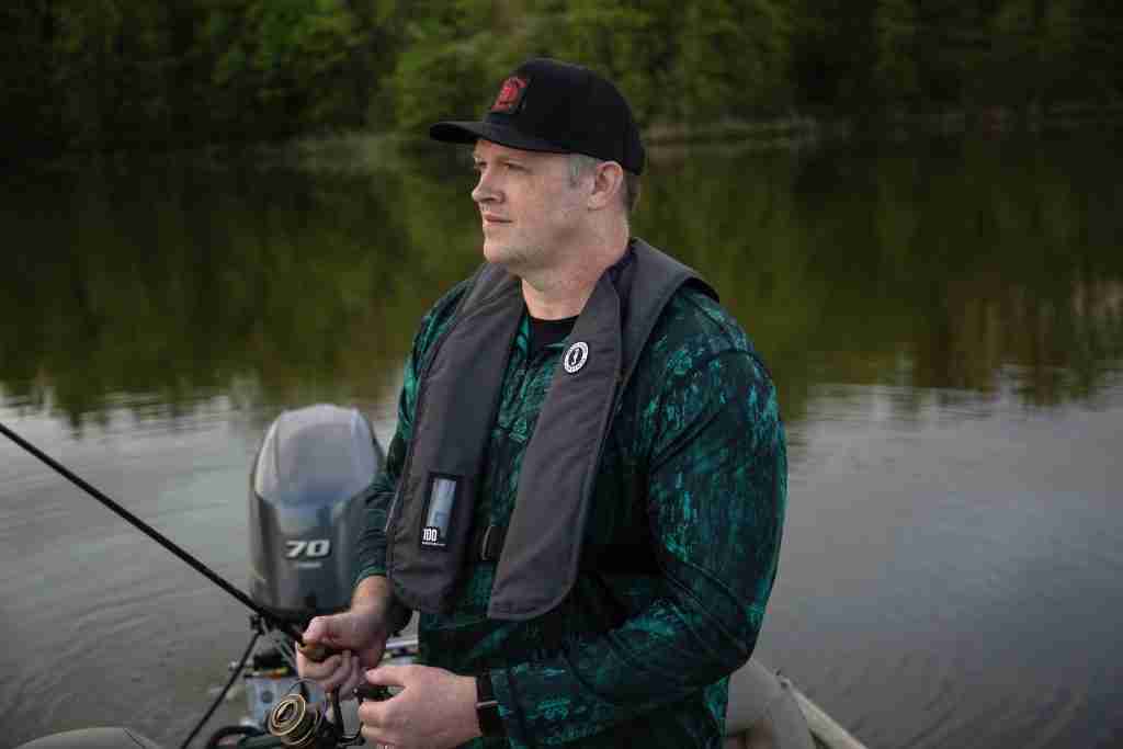 A man goes fishing in a boat on a lake while wearing an inflatable live vest