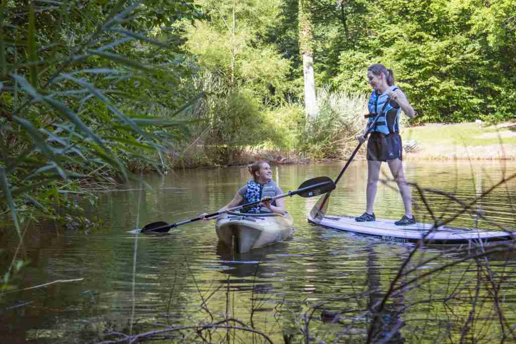 Two young people paddleboard on a lake wearing traditional foam life jackets